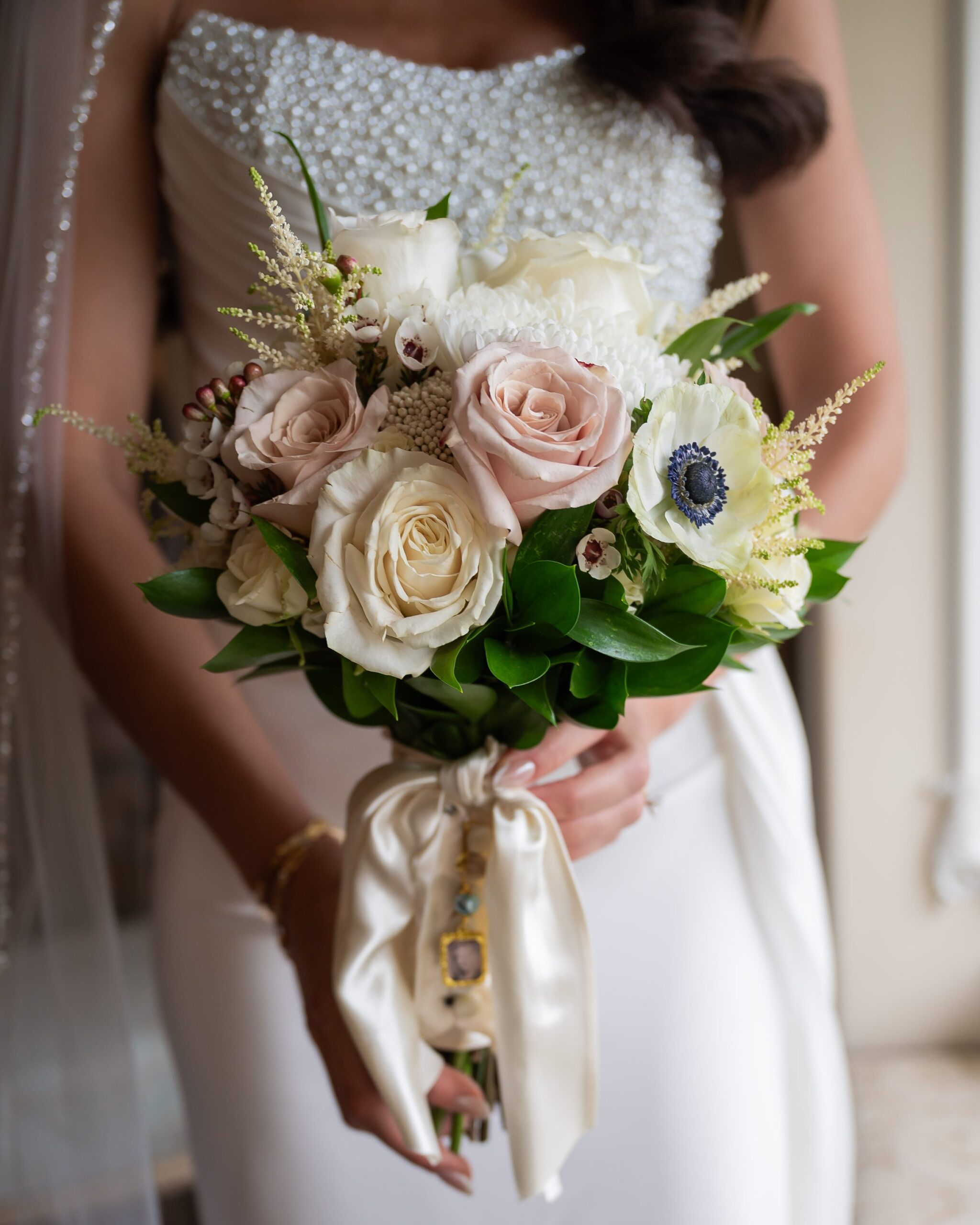 A bride holds a bouquet with white and pink roses, white anemones, greenery, and decorative ribbon on a beaded dress background.