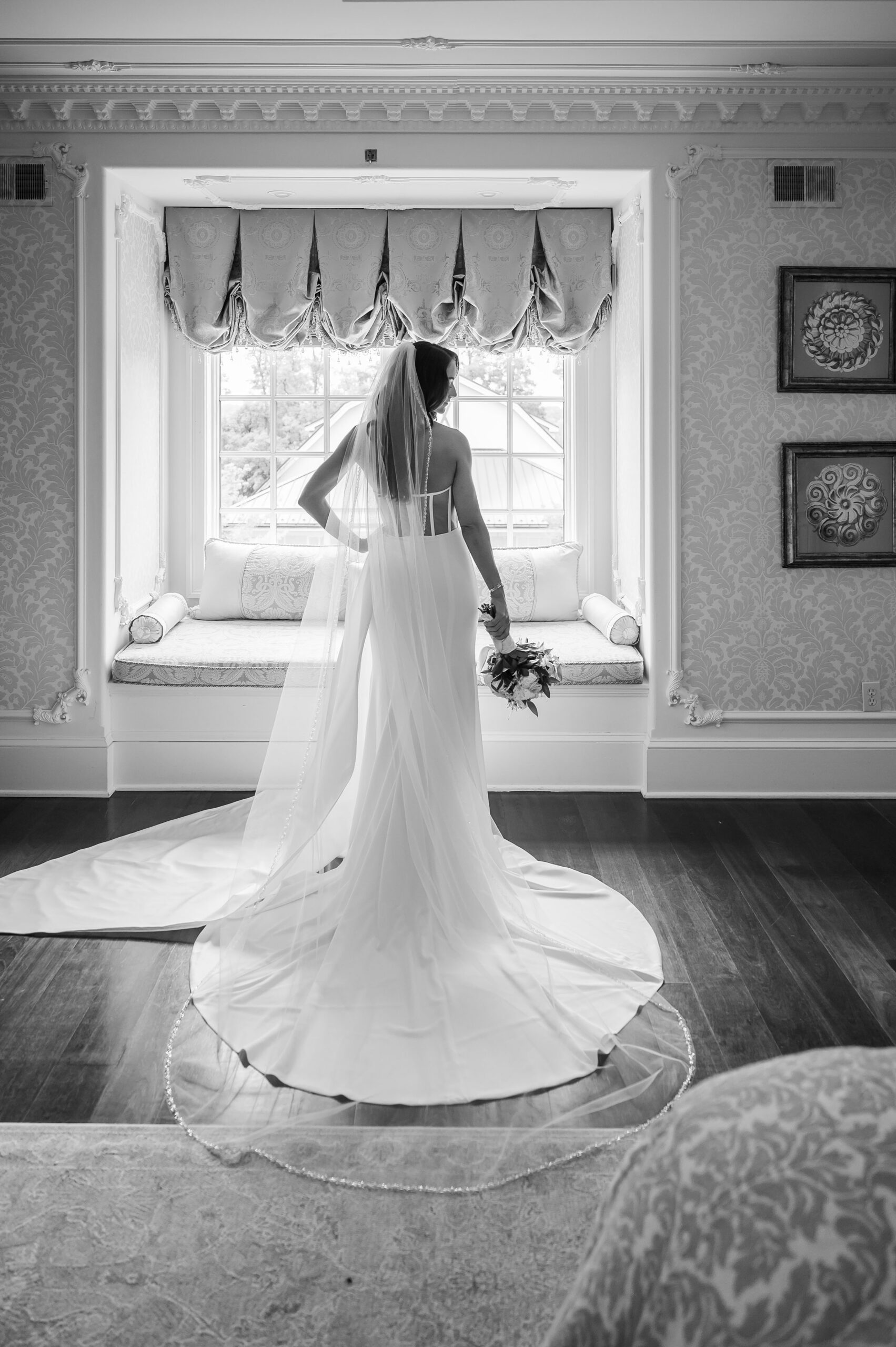 A bride in a wedding dress stands by a window, holding a bouquet, with light streaming in. The room features ornate decor and a cushioned window seat.