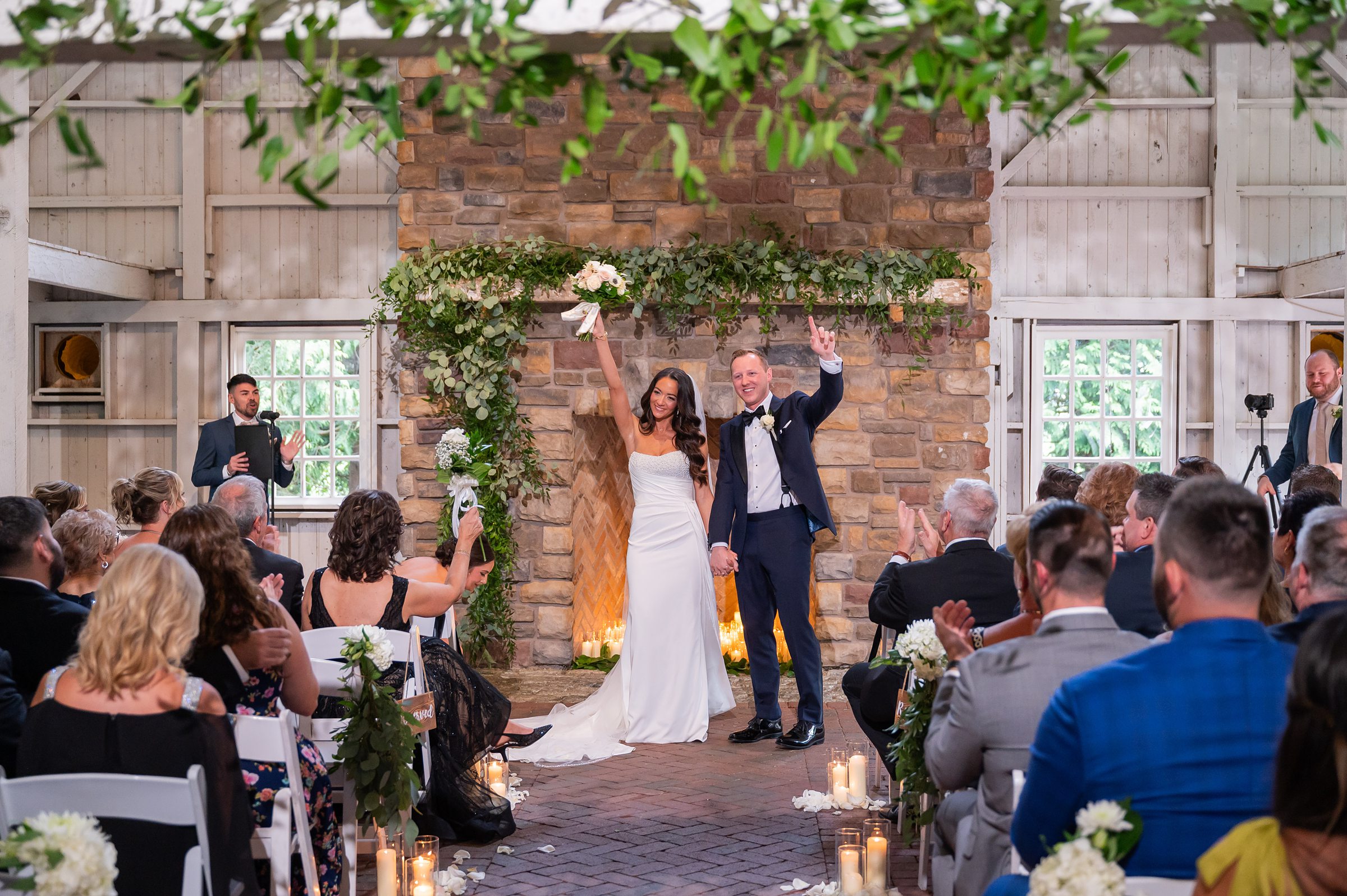 A bride and groom celebrate during their indoor wedding ceremony, standing in front of a flower-adorned stone backdrop, while guests watch and applaud.